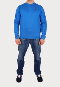 Man i en ljusblå långärmad sweatshirt, urblekta blå jeans och svarta sneakers med vita snören, står mot en vit bakgrund.