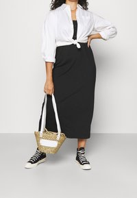 Schwarzes Maxikleid mit einem weiß gebundenen Hemd, kombiniert mit einer geflochtenen Strohtasche mit weißen Akzenten und schwarzen High-Top-Sneakern.