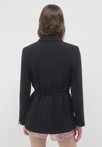 Blazer nero su misura con maniche lunghe, vita cintrata e una texture liscia. Indossato sopra pantaloni corti brillanti di paillettes rosa. Sfondo bianco uniforme.