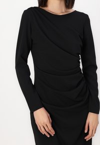 Černé dlouhé šaty s dlouhým rukávem, které mají splývavý design, hladkou texturu a lodní výstřih. Látka má jemný matný povrch.