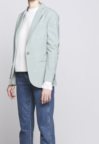 Jasnozielona dopasowana marynarka z teksturowanej tkaniny, zapinana na jeden guzik, w towarzystwie białego swetra i niebieskich dżinsów.