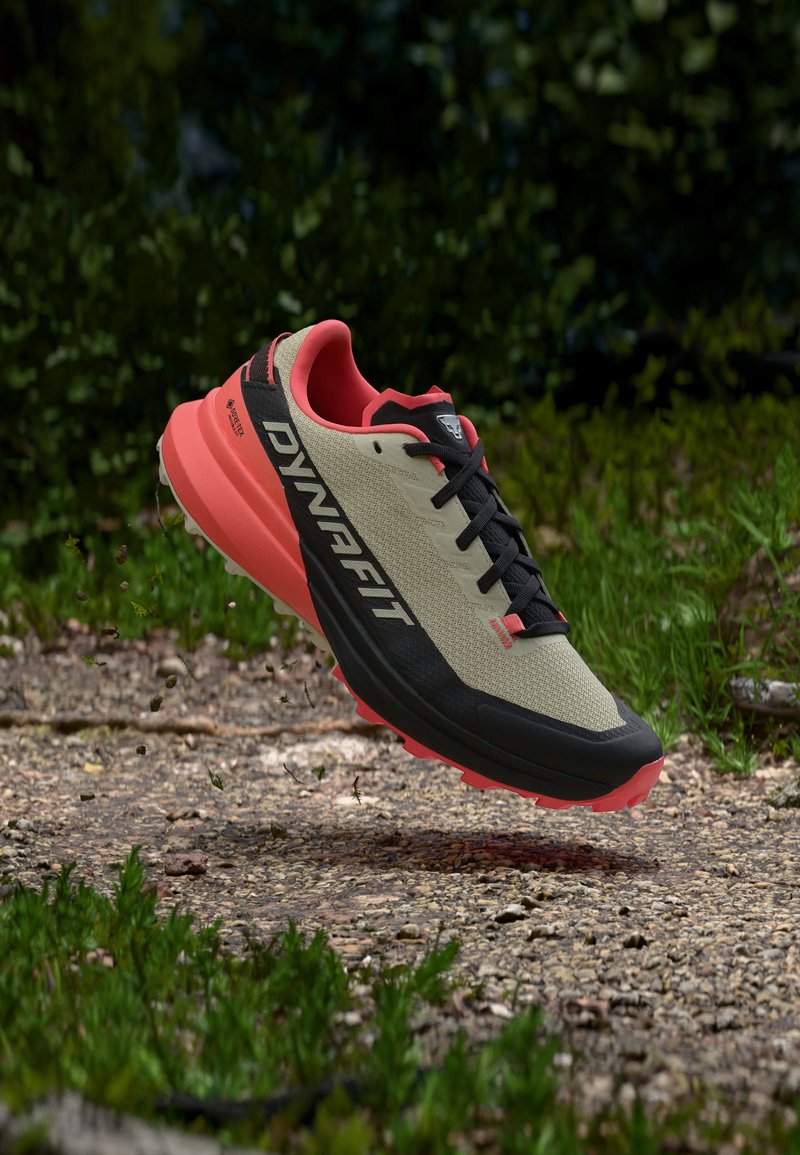 Trail running shoe with beige and coral colors, black laces, "DYNAFIT" logo on side, hovering above rocky path with greenery in background.