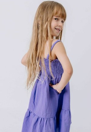 Young girl with long blonde hair wearing a sleeveless purple dress, looking over shoulder with hand on hip against plain background.