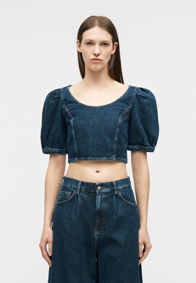 Γυναίκα με μακριά καστανά μαλλιά, που φοράει cropped μπλε τζιν μπλούζα με φουσκωτά μανίκια και τζιν παντελόνι ψηλοκάβαλο με φαρδιά πατσαβούρα.