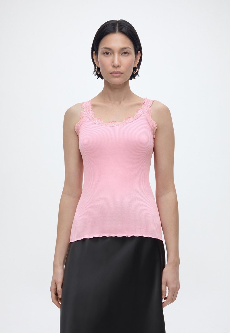 Γυναίκα με κοντά σκούρα μαλλιά, φορά ροζ πλεκτό τοπ με δαντέλα και μαύρη φούστα, στέκεται μπροστά σε μονόχρωμο ανοιχτό φόντο.