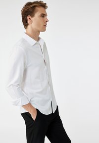 Camisa blanca de manga larga con cuello puntiagudo, cierre frontal de botones y puños enrollados. Combinada con pantalones negros, que muestran un diseño ajustado.