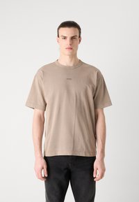T-shirt en coton beige à manches courtes et col rond ; présente un petit logo centré sur la poitrine. Porté avec un jean foncé.