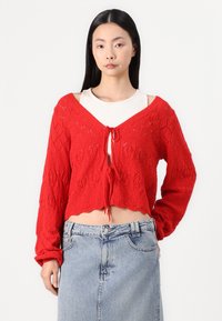 Cardigã curto tricotado em vermelho com decote em V, mangas longas e frente aberta atada com um laço. Apresenta um padrão de diamante e bainha ondulada.