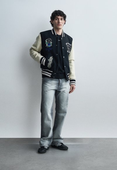 Νέος με σγουρά μαλλιά, φορώντας μαύρο και εκρού varsity μπουφάν, μαύρο πουκάμισο, ξεβαμτζιν και μαύρα αθλητικά παπούτσια, μπροστά σε απλό γκρι φόντο.