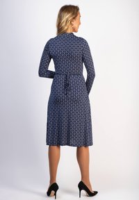 Mekko, jossa on pitkät hihat, keskipitkä leikkaus, valkoinen ympyräkuvio koko pinnalla, istuva vyötärö solmukoristeella, yhdistetty mustiin korkokenkiin.