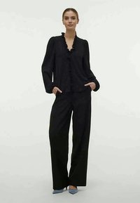 Μαύρη μπλούζα με ριγέ λαιμό και λεπτομέρειες από κουμπιά, συνδυασμένη με μαύρο παντελόνι φαρδύ και ανοιχτό μπλε τακούνια, δημιουργώντας μια συντονισμένη εμφάνιση.