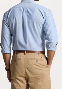 Camicia a maniche lunghe azzurro chiaro con polsini arrotolati, dettaglio della piega sul retro, abbinata a pantaloni beige con tasche e una cintura in pelle marrone.