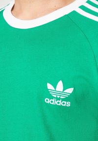 Grön bomullströja med vit ribbad krage och tre vita ränder på ärmen. Har en vit broderad Adidas-logga och ett bladdesign.