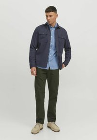 Navy button-up shirt with two chest pockets, layered over a light denim shirt, paired with olive green trousers and beige shoes.