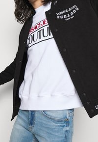 Černá vysokoškolská bunda na knoflíky s vyšívaným logem "VERSACE JEANS COUTURE". Nošená přes bílou mikinu a světlé modré džíny.