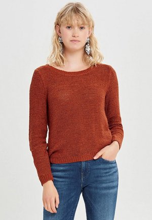 Femme portant un pull en maille couleur rouille et un jean bleu, avec une main dans la poche et des boucles d'oreilles pendantes voyantes.