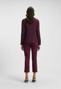 Bordeauxfarbener Blusen- und Hosenanzug. Die Bluse hat lange Ärmel und einen leicht abgerundeten Saum. Figurbetonte Hose mit cropped Länge. Schwarze High Heels.