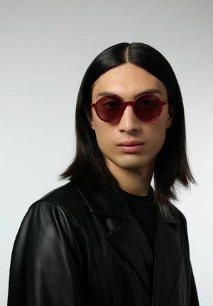 Red round sunglasses with a thin frame, worn by a model in a black leather jacket, against a light gray background.