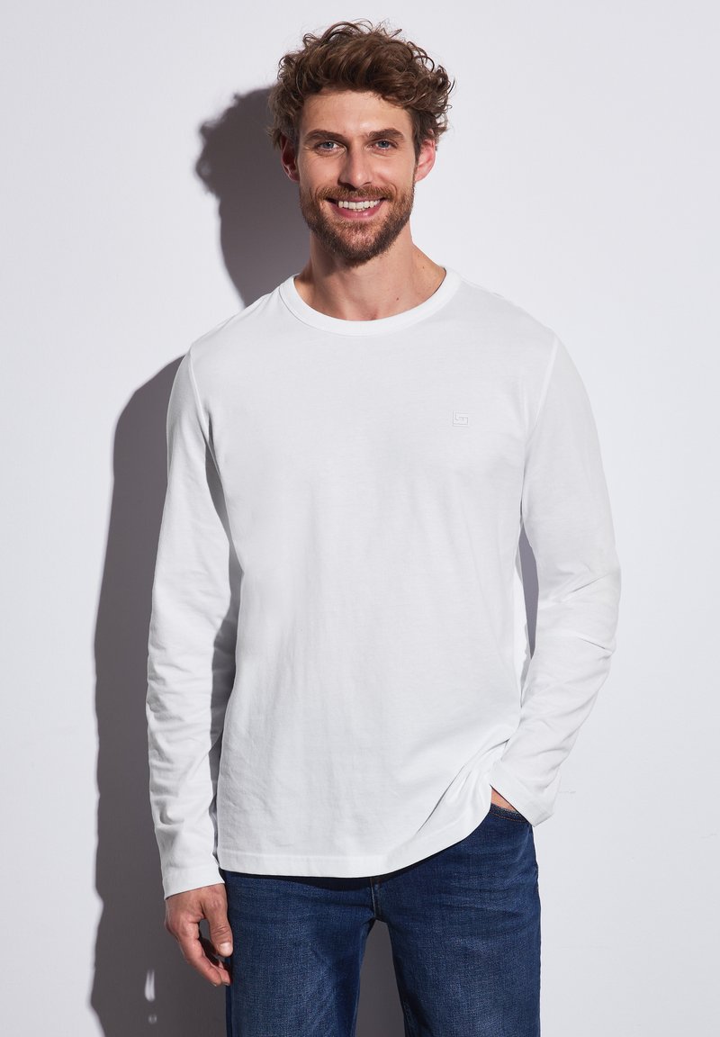Camisa blanca de manga larga hecha de algodón, con cuello redondo y un pequeño logotipo en el pecho, combinada con jeans azules.