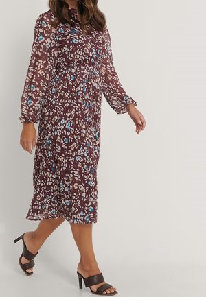 Mujer que lleva un vestido de manga larga color granate, por debajo de la rodilla, con patrones abstractos en azul, blanco y beige, combinado con sandalias negras de tacón alto.