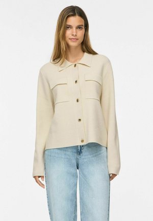 Jeune femme aux cheveux raides porte une chemise beige boutonnée avec des poches et un jean bleu clair sur un fond blanc uni.