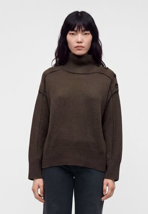 Femme aux cheveux noirs et lisses portant un pull à col roulé marron avec des boutons sur l'épaule et un pantalon foncé, debout devant un fond uni.