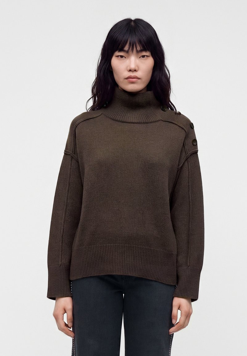 Femme aux cheveux noirs et lisses portant un pull à col roulé marron avec des boutons sur l'épaule et un pantalon foncé, debout devant un fond uni.