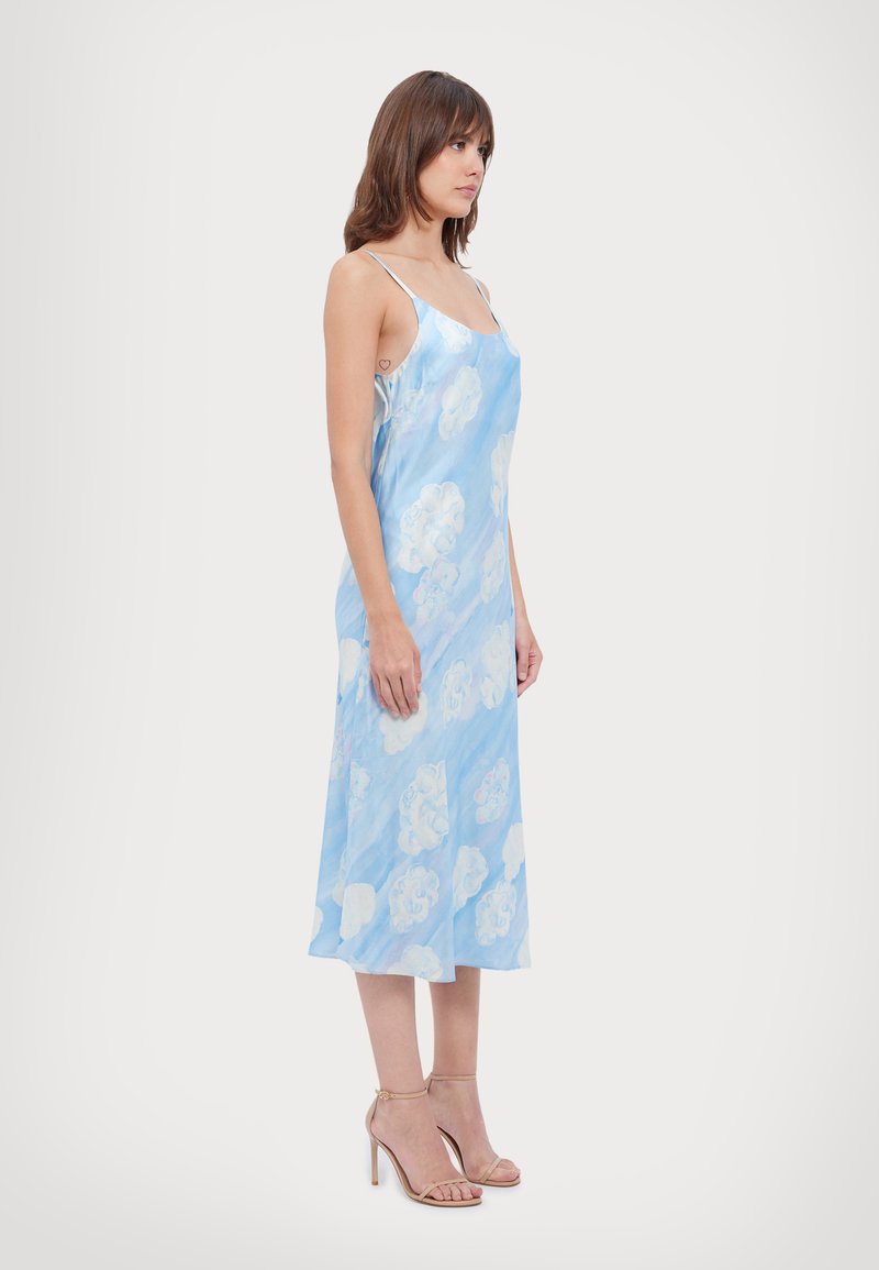 Rochie din satin de culoare albastru deschis, cu modele de nori albi, bretele subțiri și o croială lejeră care se termină la jumătatea gleznei. Asortată cu tocuri bej.