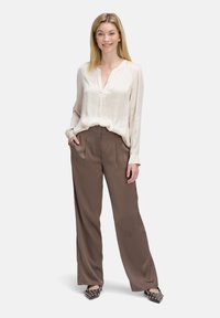 Hellbeige Seidenbluse mit partieller Knopfleiste, kombiniert mit weiten braunen Hosen. Das Model trägt dunkle Schuhe mit Muster und Nietenakzenten.