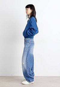 Chaqueta bomber azul combinada con jeans claros rasgados, con un corte relajado y zapatillas blancas. El fondo es liso y de color claro.