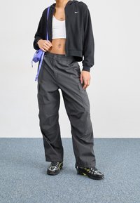 Person som bär en svart kort zip-up jacka, vit kort tanktop, grå cargobyxor, svart och grå sneakers, och bär en lila axelväska.