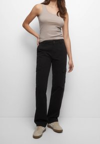 Žena v béžovém tílku, černých cargo kalhotách se bočními kapsami a béžových teniskách, stojící na jednobarevném bílém pozadí.