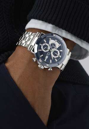 Ezüst fém kronográf óra kék számlappal és három kisebb számlappal, fehér ing mandzsetta és sötét pulóver ujja felett a csuklón viselve.