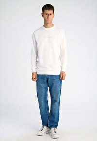 Vit sweatshirt med rund halsringning, prydd med diskret "LINDBERGH"-text, kombinerad med blå jeans och vita sneakers. Snyggt tyg.