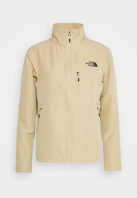 Beige mjukskaljacka med hög krage, zippered fram och två sidofickor. Har en svart logotyp på övre vänstra sidan.