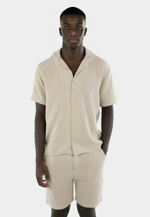 Lys beige kortermet skjorte og matchende shorts laget av teksturert stoff. Har en løs passform, knappelukking og subtilt mønster.