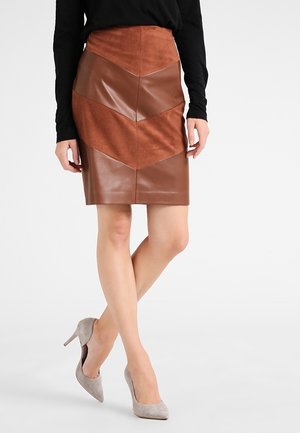 Femme portant une jupe en cuir et en daim à motifs chevrons marron, un haut noir à manches longues et des escarpins gris en daim, se tenant les jambes croisées.
