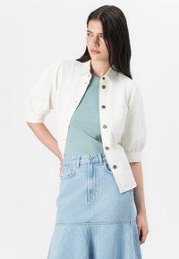 Mladá žena sa sebavedomo fotografe, oblečená v bielej oversized bunde cez tyrkysový top a svetlomodré riflové sukne. Mäkko neutrálny background.