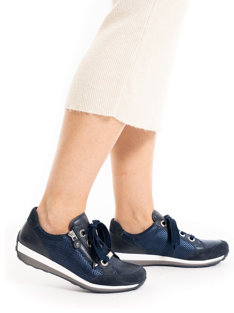 Person trägt marineblaue Sneaker mit Schnür- und Reißverschlussdetails, steht vor einem weißen Hintergrund und trägt einen cremefarbenen Rock.