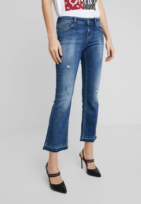 Mujer vistiendo jeans desgastados cortos de color azul y zapatos de tacón negro con tiras, de pie contra un fondo liso.