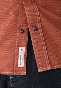 Tan textured shirt with a diamond pattern, featuring black buttons and white stitching. A fabric label displays "Durable Goods."