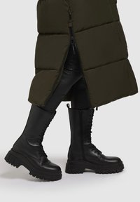 Črne vojaške čevlje odlikujejo grob podplat, vezalni del spredaj in stranski zadrga. Narejeni so iz gladke usnje z mat finišem in trpežno teksturo.