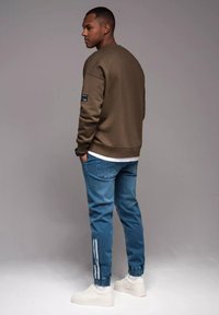 Oliwkowy sweter z ściągaczami, noszony na białej koszuli; w komplecie z jasnoniebieskimi dżinsowymi joggerami z białymi paskami i elastycznymi ściągaczami.