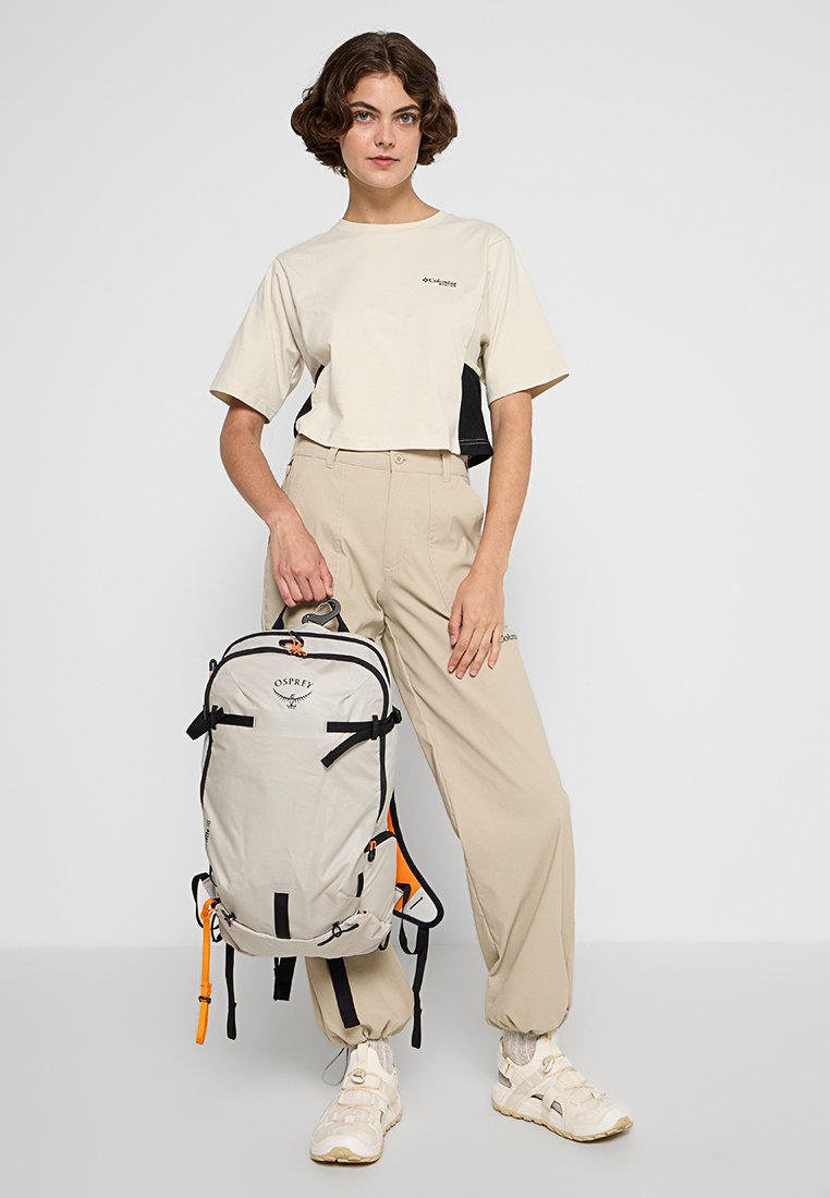 Mochila de color gris claro con detalles negros y naranjas, que cuenta con correas ajustables, un asa en la parte superior y la marca Osprey. Atuendo casual de color beige.