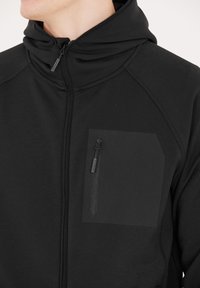 Sort zip-up hættetrøje med en tætsiddende hætte, struktureret stof og en elegant brystlomme med lynlåslukning. Glat finish og minimalistisk design.