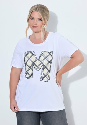 Blondynka stoi z ręką na biodrze, ubrana w biały T-shirt z dużą wzorzystą literą M i szare spodnie na białym tle.