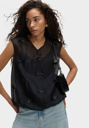 Schwarzes ärmelloses Top aus durchscheinendem Stoff, mit V-Ausschnitt und mehreren kleinen Textil-Schleifen. Kombiniert mit hellen, verblassten Denim-Hosen.