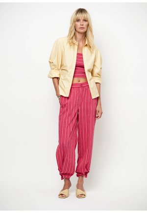 Femme portant une chemise jaune boutonnée sur un crop top rayé rouge, un pantalon rayé rouge avec des poignets noués, et des chaussures tissées beiges sans lacets.