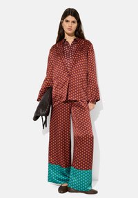 Bluzka satynowa w kropki oraz szerokie spodnie w kolorze burgundowym z pomarańczowymi kropkami, z turkusowymi akcentami na brzegu. Modelka trzyma czarną torbę.