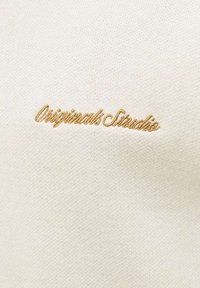 Tissu couleur crème avec broderie dorée affichant en cursive le texte "Originals Studio" centré sur le tissu.